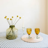 A styled tabletop scene with a translucent green glass vase holding yellow dried flower stems, two amber glass goblets on a round woven placemat, and a white candle in a clear holder, all set on a green-and-white striped tablecloth against a warm off-white wall. All made in Mexico.
