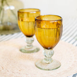 Two amber glass goblets with clear glass bases, arranged on a woven beige placemat with a striped tablecloth and a blurred green glass vase in the background.