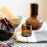 Chocolate Brown Round Glass Carafe with lid and a matching chocolate brown glass tumbler styled on a striped cloth with dried red Mexican chili peppers in a dark bowl.