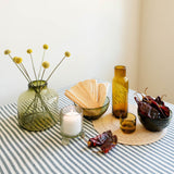 A styled tabletop arrangement on a hunter green-and-white striped cloth, featuring a green glass vase with dried yellow flowers, a white candle in a glass holder, a small amber bottle carafe, a bowl of dried red peppers, and other decorative glassware against a plain cream wall.