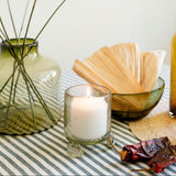 A styled tabletop scene with a lit white candle in a clear glass holder, a green glass vase holding dried yellow flowers, a bowl of dried corn husks, and a striped fabric runner against a soft off-white background.