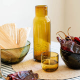 An amber glass carafe bottle and matching small tumbler that serves as lid on a striped tablecloth, styled with dried red chilies in a dark bowl and corn husks in a green bowl against a soft beige background.