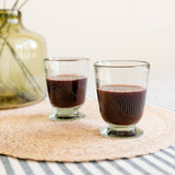 Two clear handblown glass cups filled with wine, placed on a woven round placemat with a striped tablecloth and a blurred green glass vase in the background.
