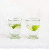 Two clear Mexican handblown glass tumblers with footed bases, each containing clear water and a lime wedge, set against a plain white background.