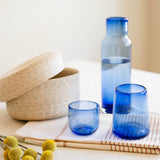 A set of translucent cobalt blue glassware, including a tall carafe, a tumbler, and a small tumbler, arranged on a striped napkin next to a woven beige basket and yellow pom-pom flowers.