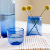 A set of cobalt blue glass tableware, including a tall bottle, a small tumbler, and a cylindrical vase holding yellow pom-pom flowers, arranged on a beige striped cloth on a light surface.