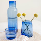 Blue glass home decor set including a tall bottle, a short round  tumbler, and a cylindrical vase with dried yellow flowers, arranged on a woven placemat with a striped napkin against a warm off-white background.