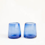 Two small cobalt blue handblown glass tumblers with a gradient fade from translucent blue at the top to darker blue at the base, set against a plain white background.