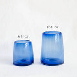 Two translucent cobalt blue hand blown glass tumblers of different heights, standing on a white textured surface against a white background.