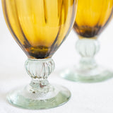 Two amber glass goblets with flared bases and ribbed stems, photographed on a white surface with shallow depth of field.