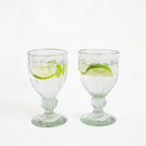 Two clear handblown glass goblets with flared bases, each containing water and a lime slice, photographed against a plain white background.