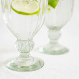 Two clear ribbed handblown glass goblets with pedestal bases on a white surface, each holding a lime slice garnish, photographed in soft natural light.