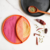 A round woven cloth cotton tortilla warmer with pink and orange color block design, shown partially open with a stack of tortillas inside. It is styled on a white marble surface with a black plate, a wooden spoon of pumpkin seeds, and dried red chilies.
