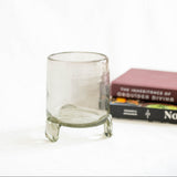 A clear handblown glass candle holder with a cylindrical body and three short legs, shown on a white surface with two books blurred in the background.