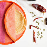 A close-up of a cloth pink and orange tortilla warmer with tortillas inside styled with dried red chilies, green pumpkin seeds, and a wooden spoon of seeds on a dark plate over a white marble surface.