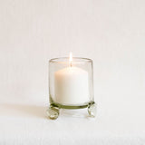 A lit white pillar candle in a clear glass holder with a small footed base, photographed against a soft white background.