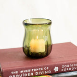 An olive green handblown glass candle holder containing a lit candle, resting on top of a brown book against a plain white background.