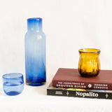 Three glass vessels in blue and amber tones arranged on a white surface with two books behind them. An amber petal fluted hurricane with a votive candle inside.