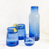 A set of cobalt blue hand blown Mexican glassware including a tall bottle carafe, a small round tumbler, and a set of two tapered glass tumblers, arranged on a white surface against a white background.