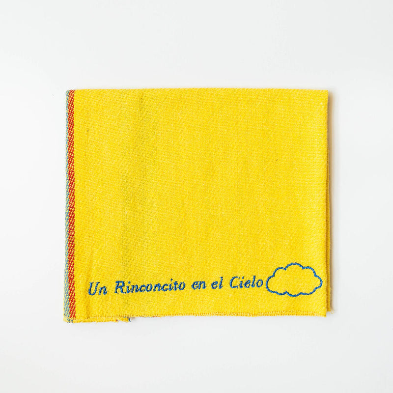 A close up of a yellow recycled cotton cleaning cloth known as Jerga with blue embroidery reading "Un Rinconcito del cielo" and a blue cloud