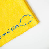A close up of a yellow recycled cotton cleaning cloth known as Jerga with blue embroidery reading "Un Rinconcito del cielo" and a blue cloud