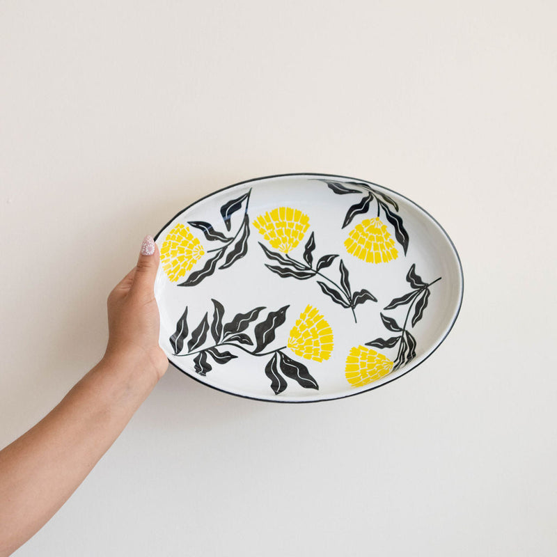 Hand holding a yellow Enamel Peltre serving tray in white with delicate orange floral accents inspired by the cempasúchil flower. The tray features a smooth, glossy finish and raised edges for easy carrying, perfect for serving snacks, or as a decorative centerpiece.