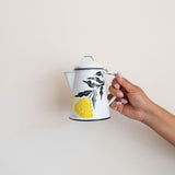 A hand holding a white enamel (peltre) kettle with vibrant yellow marigold designs and black leaf accents, set against a wall.