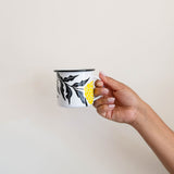 Hand holding a White enamel (Peltre) Cup with vibrant yellow marigold (cempasúchil) designs and black leaf accents, set against a wall.