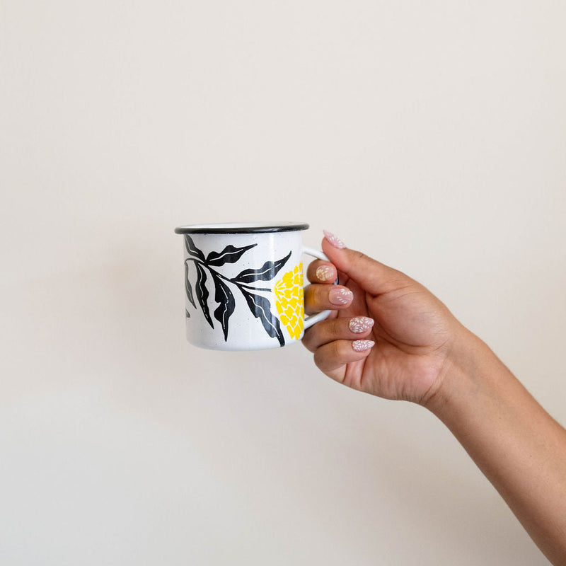 Hand holding a White enamel (Peltre) Cup with vibrant yellow marigold (cempasúchil) designs and black leaf accents, set against a wall.