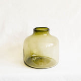 A large rounded glass vase with a short neck, made of translucent olive green glass, set against a plain white fabric background.