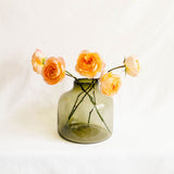 A translucent olive-green glass vase holding several peach and orange roses, photographed against a soft white fabric backdrop.