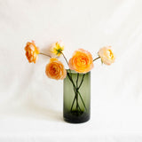 An olive green glass vase holding several orange and peach real roses, against a soft white fabric backdrop.