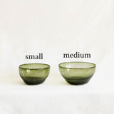 Two translucent olive green handblown glass bowls on a white fabric surface against a white background.