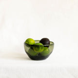 A translucent olive green glass bowl filled with limes and an avocado, set against a plain white fabric background.