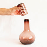 Hand holding a glass amethyst cup to an amethyst round glass carafe against a white background