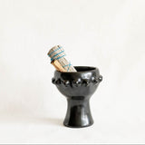 A black ceramic candle holder or incense holder with a flared rim and decorative protruding spikes, holding a bundled dried herb or smudge stick wrapped in blue string, photographed against a plain off-white background.