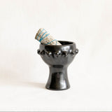 A black ceramic incense or candle holder with a flared rim, with a bundle of sage, and a short pedestal base, photographed against a plain off-white background.
