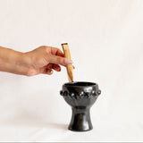 A hand holding a piece of copal over a black ceramic incense or candle holder with a flared rim and a short pedestal base, photographed against a plain off-white background.