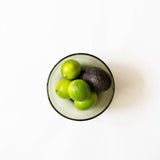 A top-down view of a round handblown olive green glass bowl containing three green limes and one dark avocado on a clean white background.