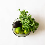 A top-down view of fresh ingredients in an olive green glass bowl on a white background, including cilantro, an avocado, and limes.