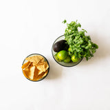 Two olive green handblown glass bowls on a white background, one containing tortilla chips and the other containing cilantro, lime, and an avocado.