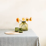 Handblown glass vse with flowers on a tablecloth-covered table against a plain wall and two olive green tumblers