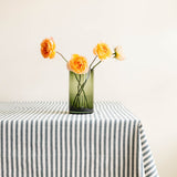 A green cylinder glass vase holding four orange roses, placed on a table covered with a green and white striped tablecloth against a plain beige wall.