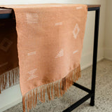 A terracotta-colored woven cotton table runner with white geometric patterns and fringe, draped over a black metal table in a minimalist room setting.