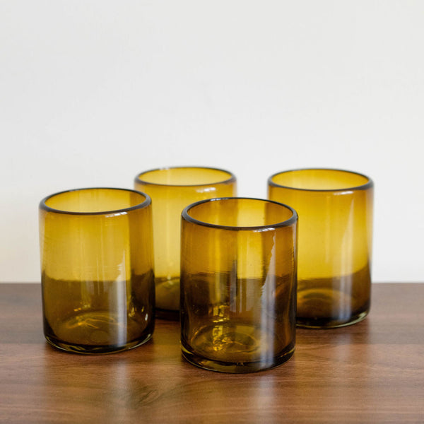 Four amber-colored Mexican recycled glass tumblers on a wooden surface with a white background