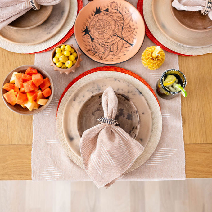The wooden rings are holding brown sacred lines napkins with subtle white geometric patterns. They're placed on earthy ceramic plates with a rustic finish that have reddish-orange rims. The entire arrangement sits on a light wooden table with a minimalist, natural aesthetic.