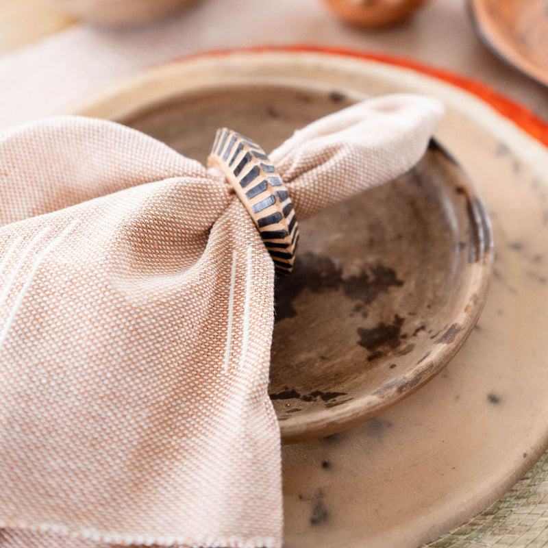 The wooden rings are holding brown sacred lines napkins with subtle white geometric patterns. They're placed on earthy ceramic plates with a rustic finish that have reddish-orange rims. The entire arrangement sits on a light wooden table with a minimalist, natural aesthetic.