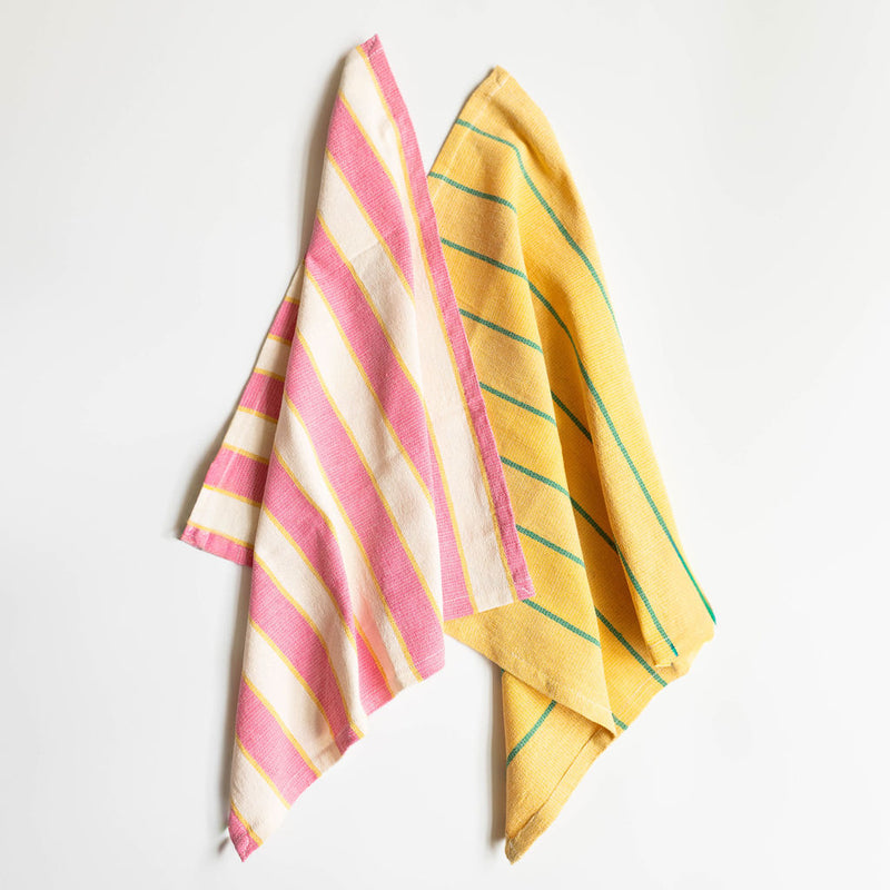 Two folded cotton napkins arranged on a white background. The top napkin features cream/off-white fabric with wide pink stripes and thin yellow accent lines. The bottom napkin displays a bright yellow fabric with thin green horizontal stripes. Both textiles showcase traditional Mexican cotton weaving techniques with vibrant colors and striped patterns