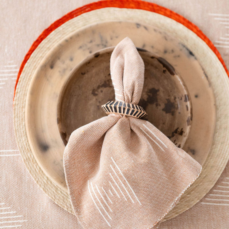 The wooden rings are holding brown sacred lines napkins with subtle white geometric patterns. They're placed on earthy ceramic plates with a rustic finish that have reddish-orange rims. The entire arrangement sits on a light wooden table with a minimalist, natural aesthetic.