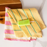 two colorful textiles on a marble countertop with a wooden cutting board. The top textile is yellow with green vertical stripes, while the bottom one features pink and white stripes with yellow accents. There's a lime green limon popsicle on a stick resting on top of the yellow textile.